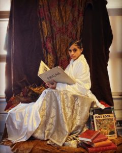 A girl sits dressed all in white reading. There are books on the ground beside her and a dramatic curtain in the backdrop