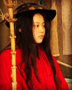 A girl looks off to the right. She has long black hair and is wearing a hat and a red shirt. There is a bamboo cane standing beside her.