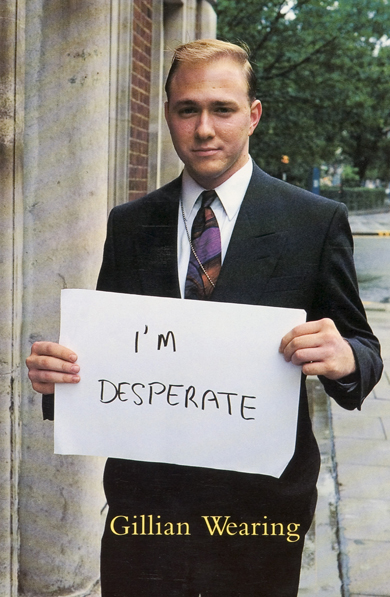 A man stands on the street wearing a suit. He holds up a handwritten sign that says 'I'm Desperate'