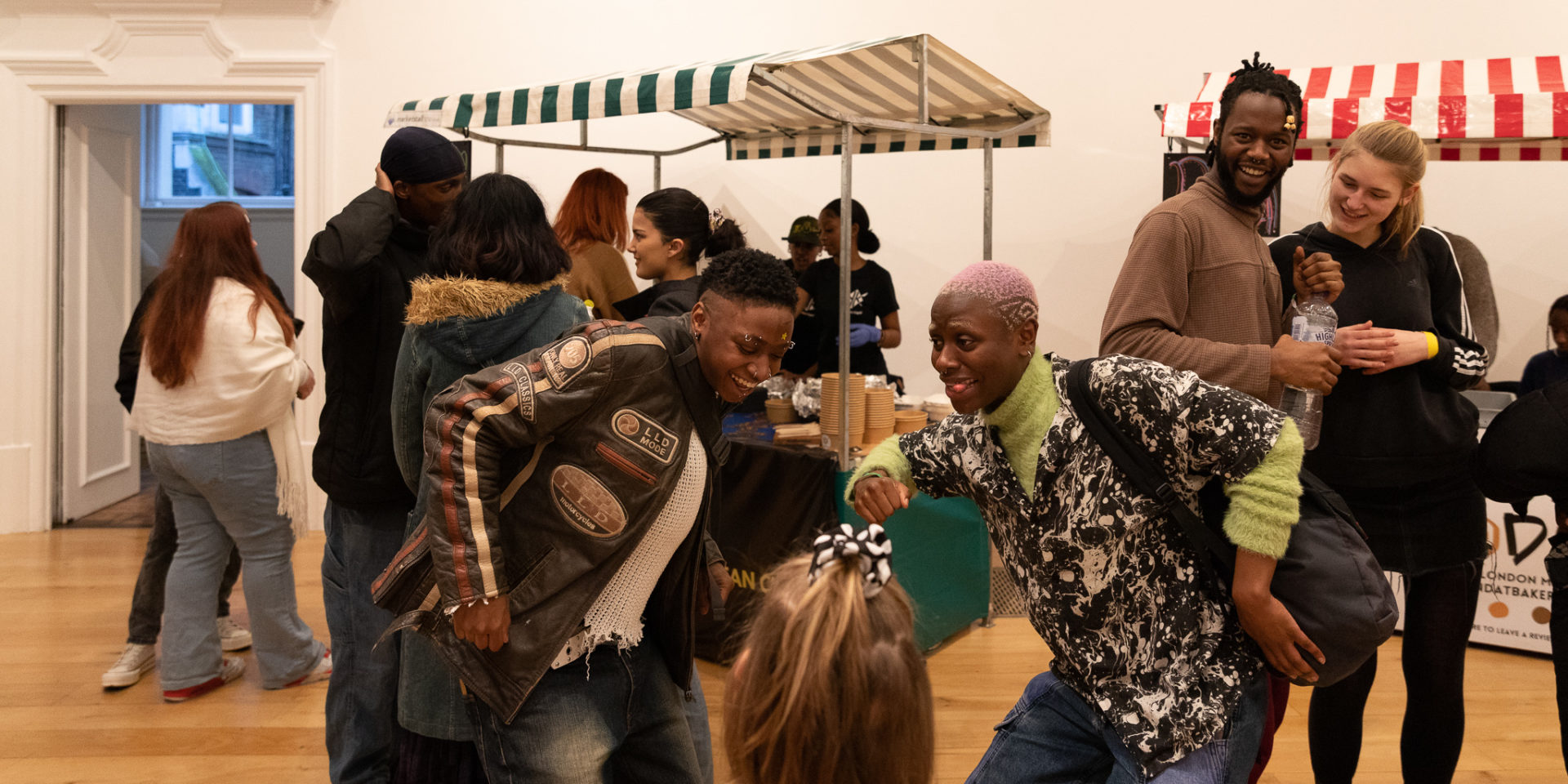 Two young people dance together beside a young child. Other people watch them and walk around. There are market stalls behind them.