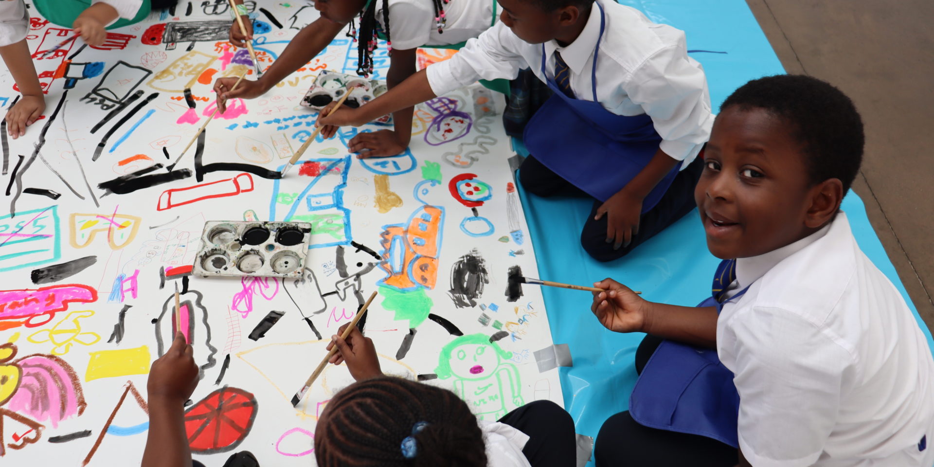 A young child sits on a bright blue mat on the floor looks at the camera. He is in a school uniform and is sitting beside other school children. The children hold paintbrushes and are all painting in bright colours onto a white canvas laid on the floor.