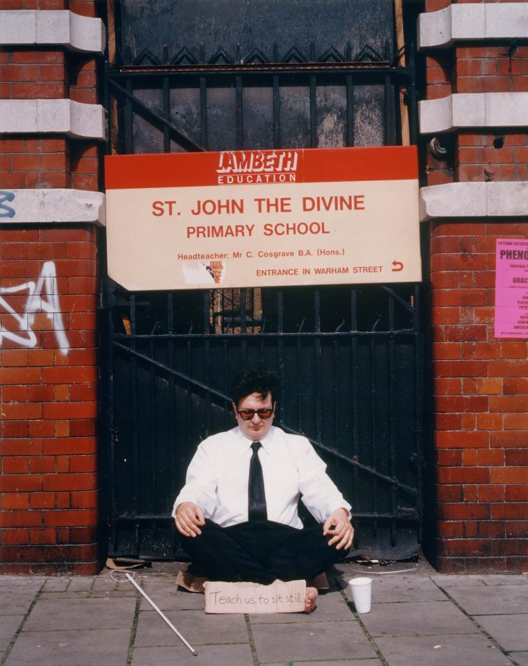 A man wearing a suit sits on the ground in front of a primary school gate.