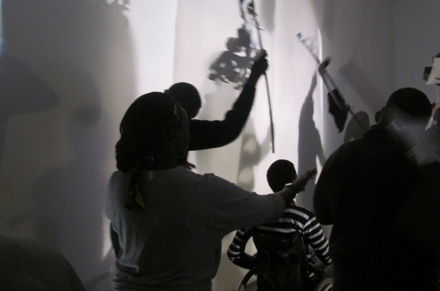 This is a black and white image of several figures including a wheelchair-user facing away from the camera towards a brightly lit wall. The group are creating artful shadows on the wall with paper creations on sticks.