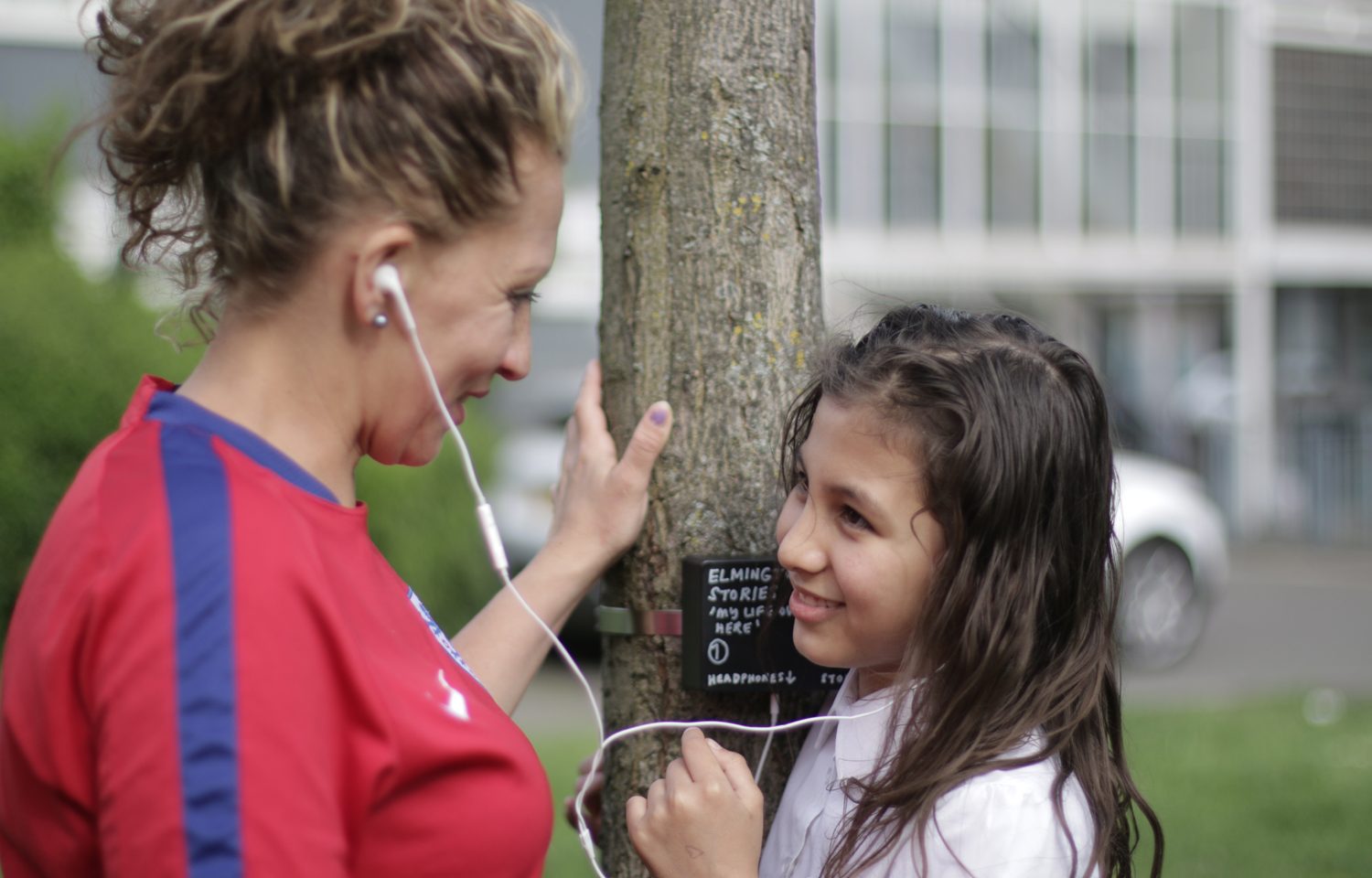 A woman and a girl are looking at each other and smiling. They are each listening to one headphone, and both are touching a tree in the middle of them.