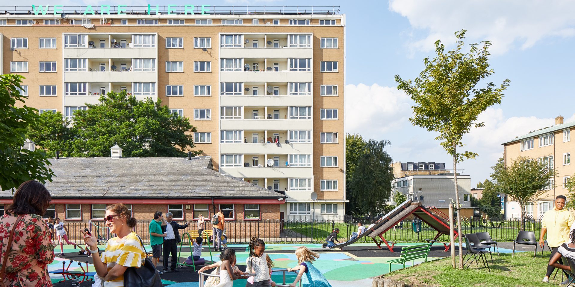 Text work 'WE ARE HERE' on top of block of flats; children playing in playground, with brightly painted surface