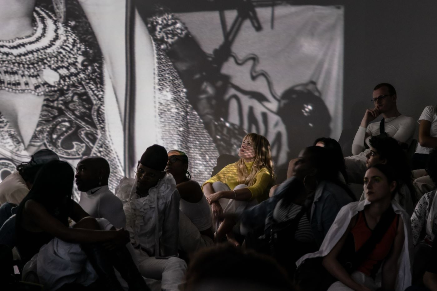 People sitting down in a dark room. There are white light projections on the walls. A woman in yellow is being illuminated by the projections.