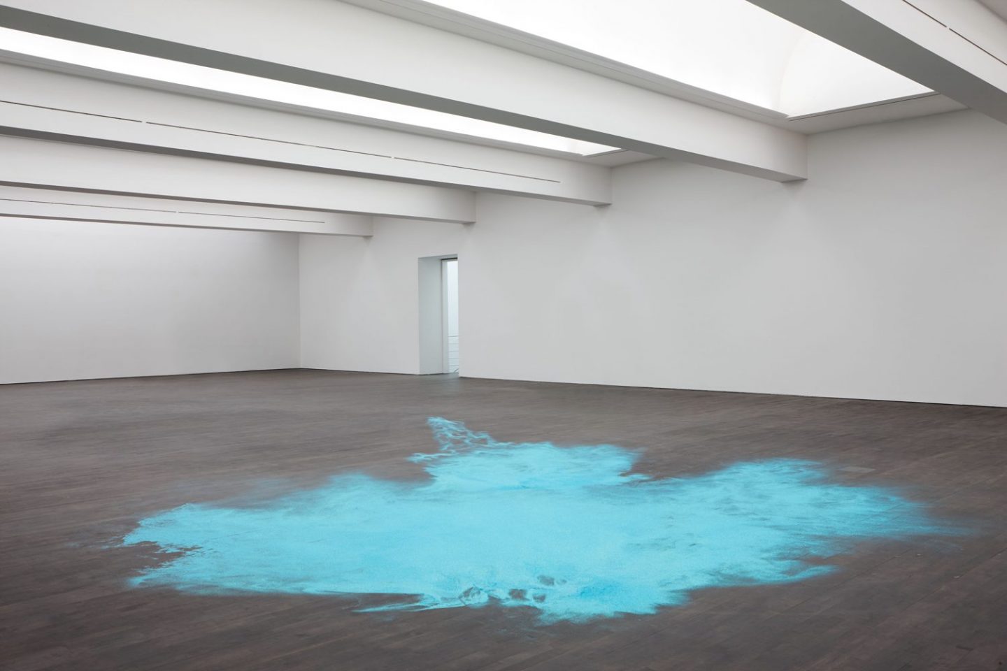 An expanse of bright blue glitter on the floor in a gallery space with white walls and grey concrete floor