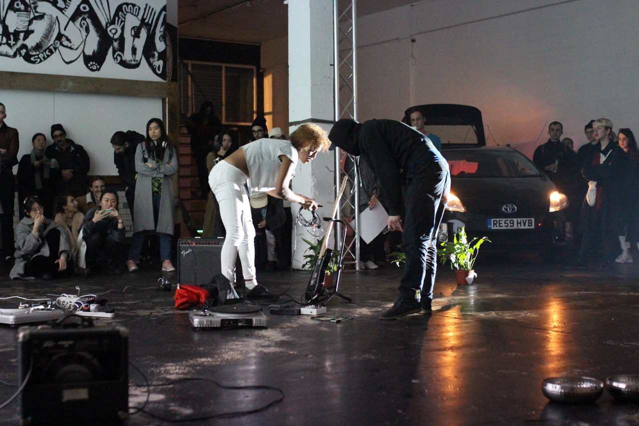 Two people stand in the centre of a room, one person is holding recording equipment and there is a speaker, turntable and houseplants on the floor. An audience watches them from the side-lines