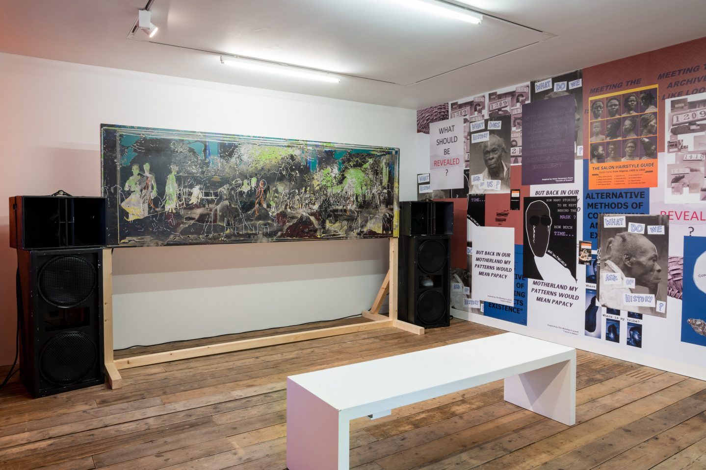A series of posters on the right hand wall, a collage painting on a wooden frame and two huge speakers on either side. A white bench is also seen positioned facing the wooden frame.
