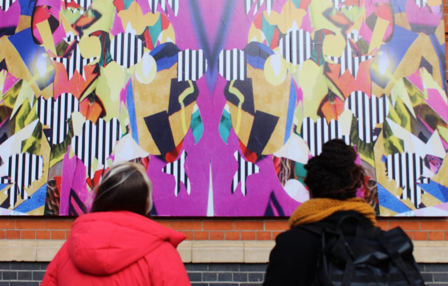 Two people wearing coats look up at a billboard of brightly coloured patterns