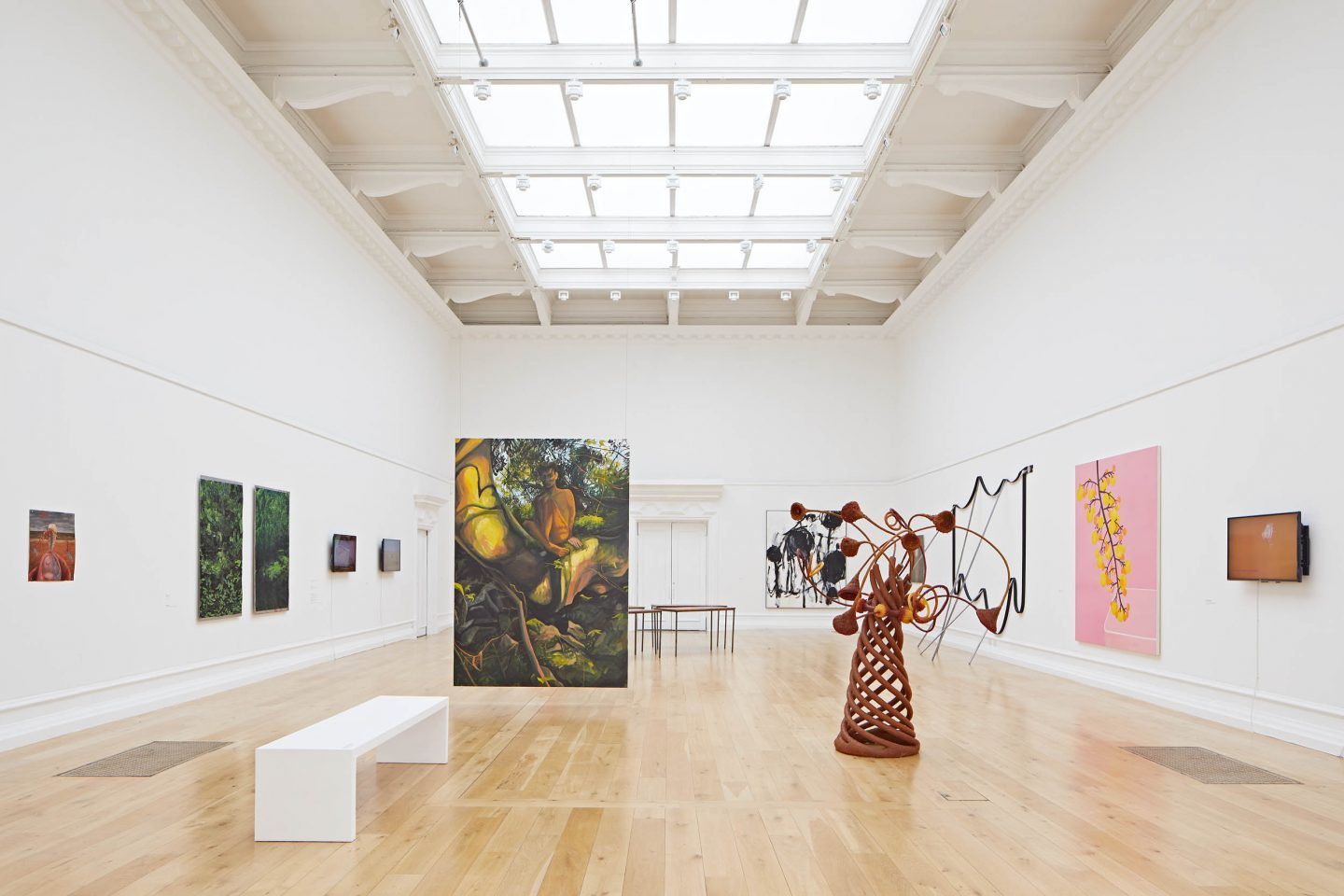 The SLG's Main Gallery with paintings on the wall, a sculpture with a conical shape and tree like limbs protruding out, and a painting hangs from the ceiling in the middle of the room