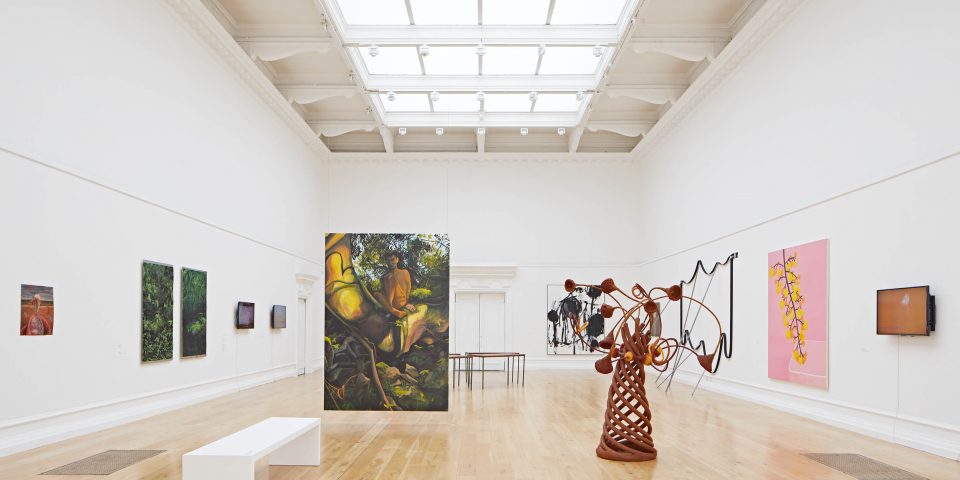 The SLG's Main Gallery with paintings on the wall, a sculpture with a conical shape and tree like limbs protruding out, and a painting hangs from the ceiling in the middle of the room