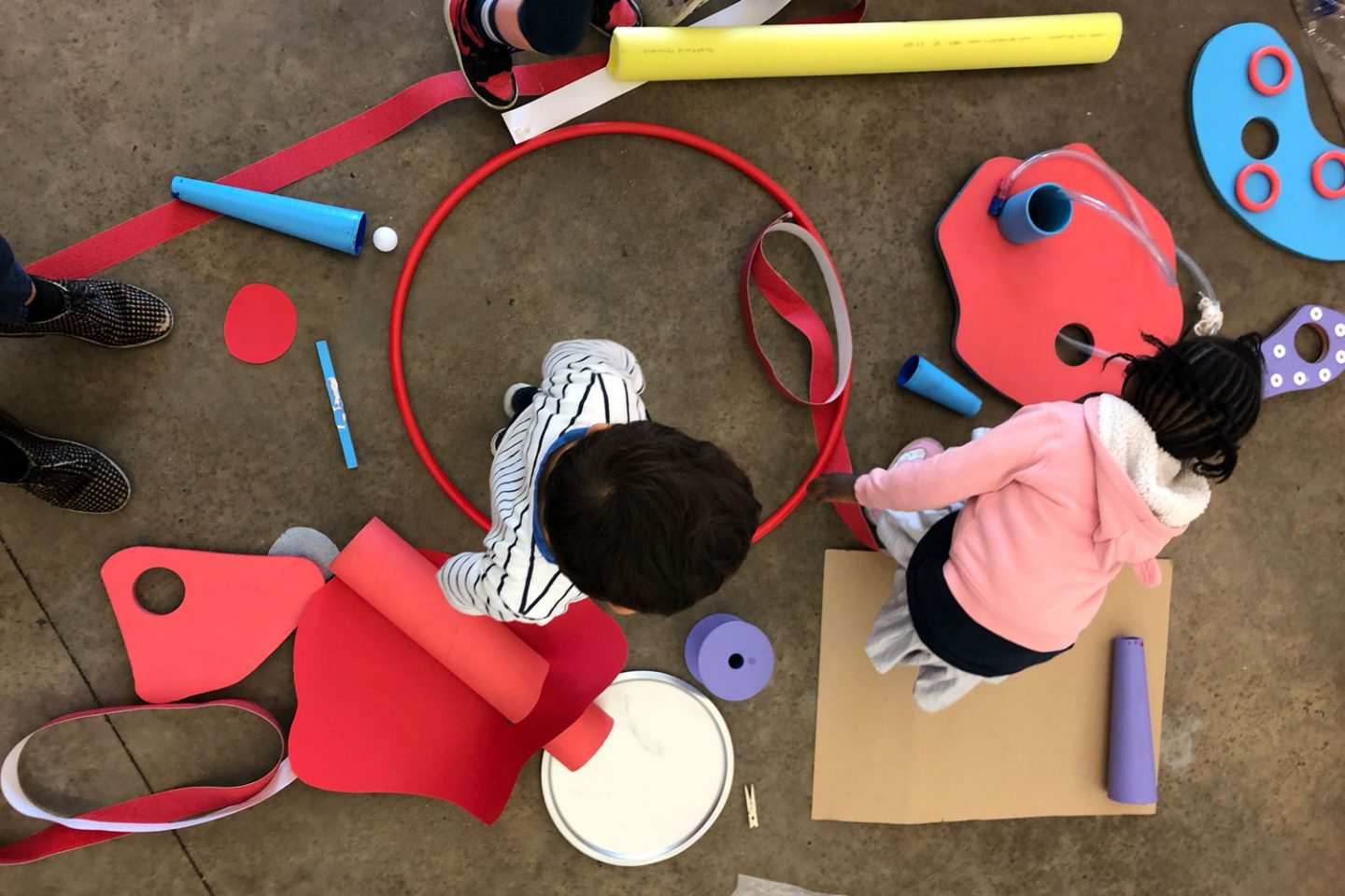 Two children surrounded by colourful paper, a hula hoop, card, and tape