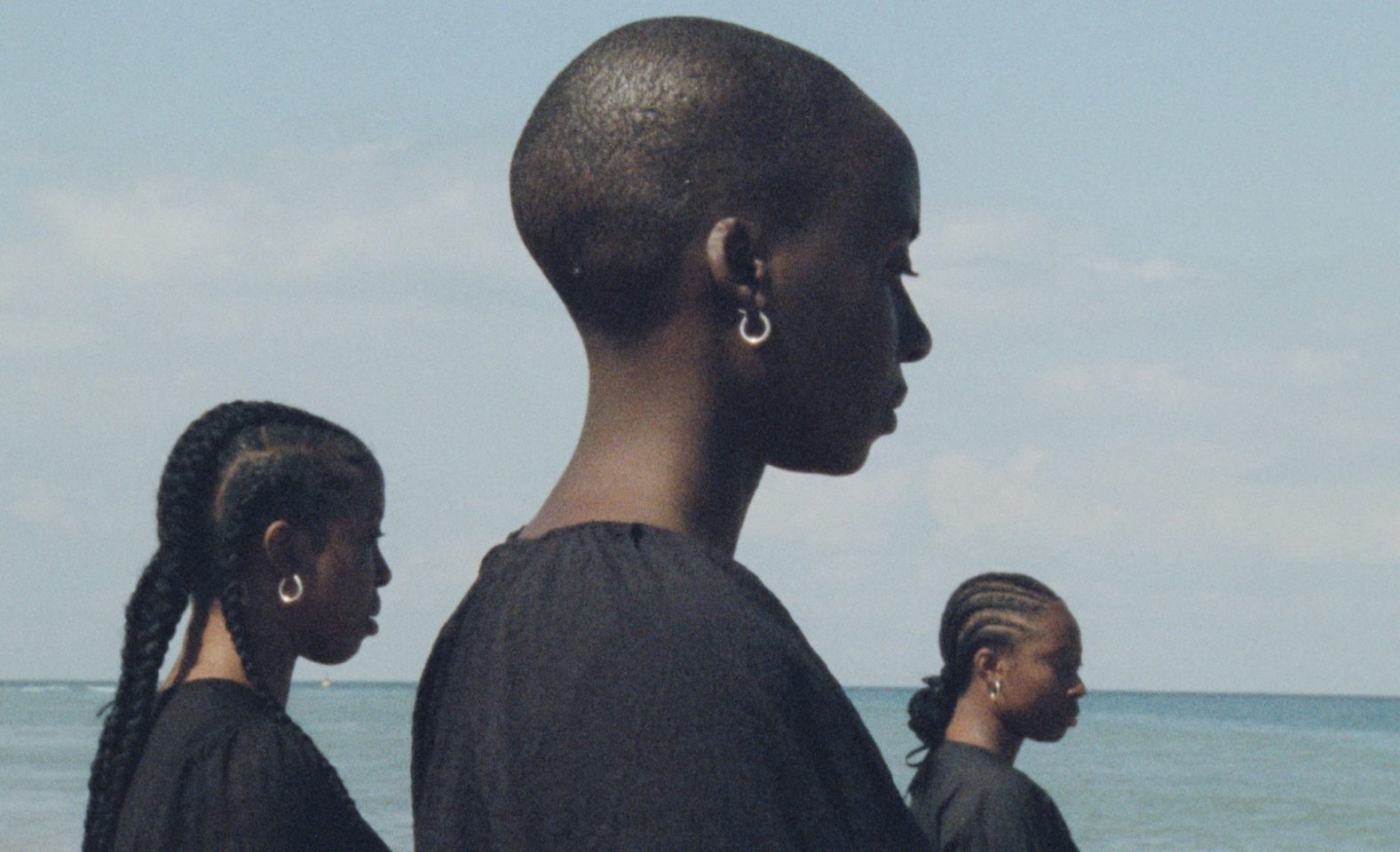 A photo of three people looking to the right, they are wearing black tops and hooped jewellery. The middle person in closest to the camera, then the person on the left is second closest and the person on the right is the furthest away. They are stood by the coast as the sea can be seen behind them.