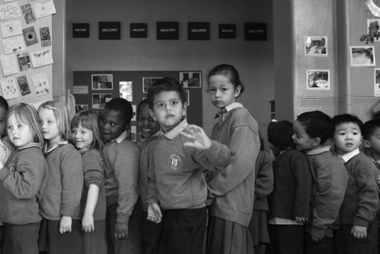 A black and white photograph of young children in school uniforms who are stood in a line, looking in various directions with a mixture of facial expressions