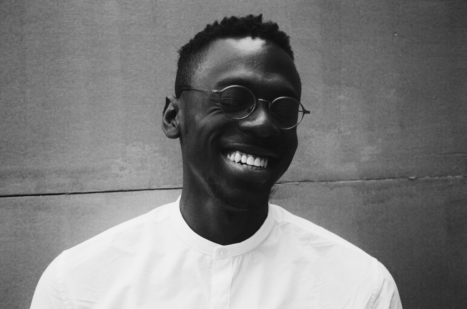 Black and white photo of Axel Kacoutié who wears glasses and is smiling with his eyes closed