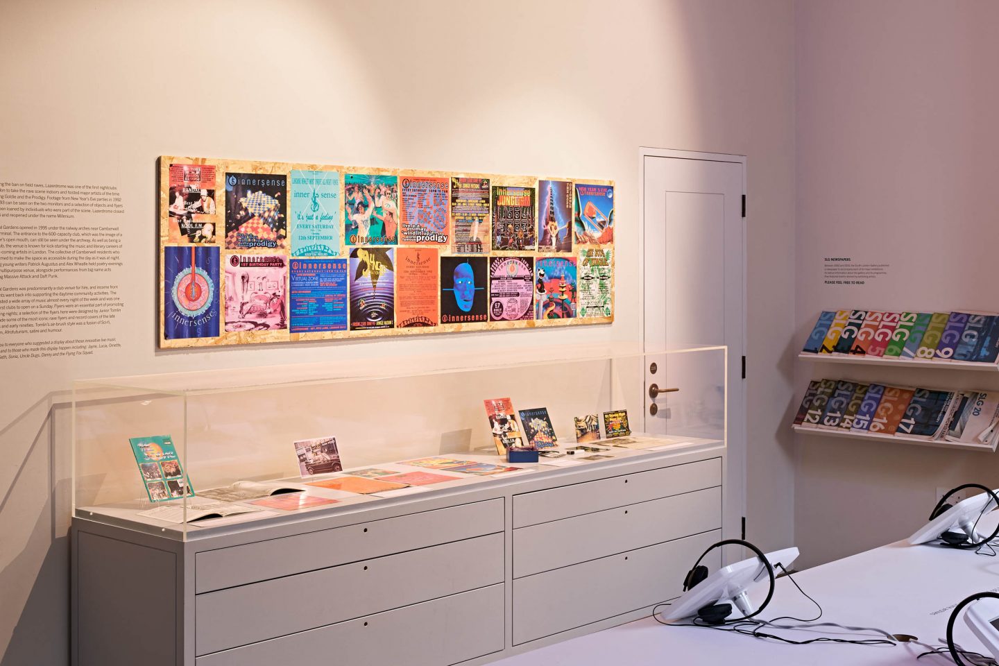 A photo of a gallery space with a display case and a series of posters on top of it. There are also two shelves in the backof the room on the right-hand side and a table with three headphones placed on top.