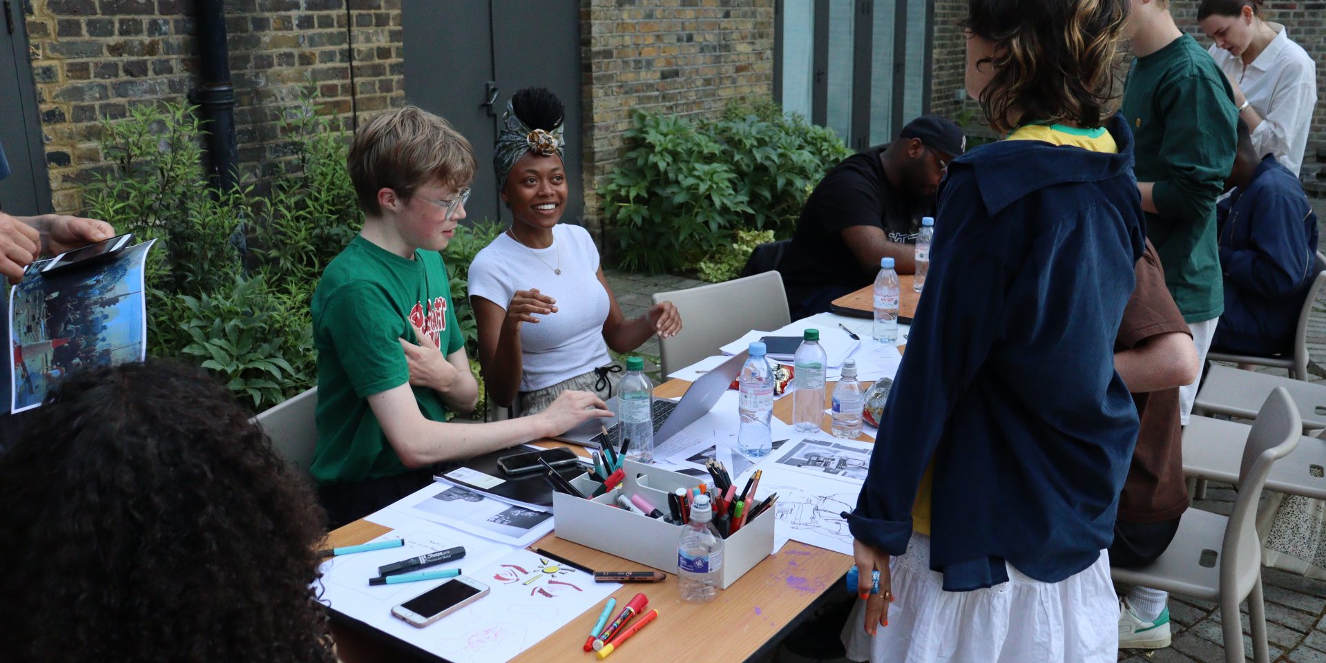 A group of young people are working around a table, smiling and chatting whilst they work on producing their zine