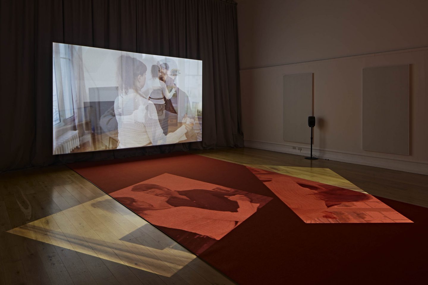 A photo of a gallery space with a large screen in front of large curtains can be seen with a speaker on the right side of the gallery space. There is a narrow red carpet in the middle of the gallery leading to the screen. There are two diagonal projections of the same film shown on the screen on the floor of the gallery space. The screen is showing a film with the artist (Alice Theobald) on the left and a man on the right.