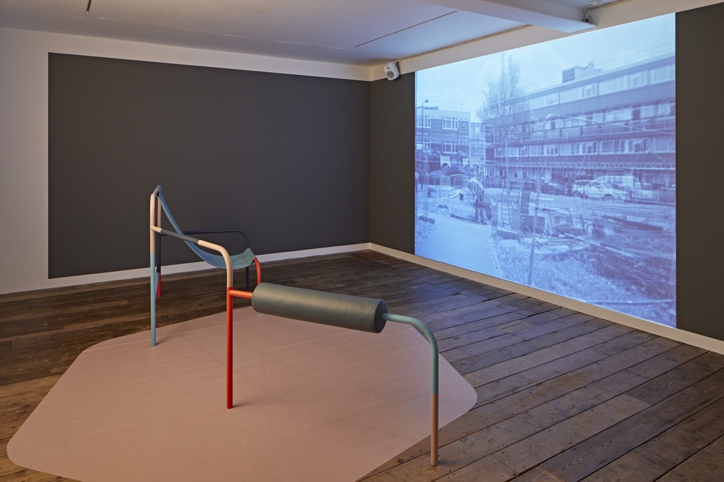 An image of a dark gallery space with a playground structure in the centre of a room facing a screen. The screen is showing a film of a housing estate.