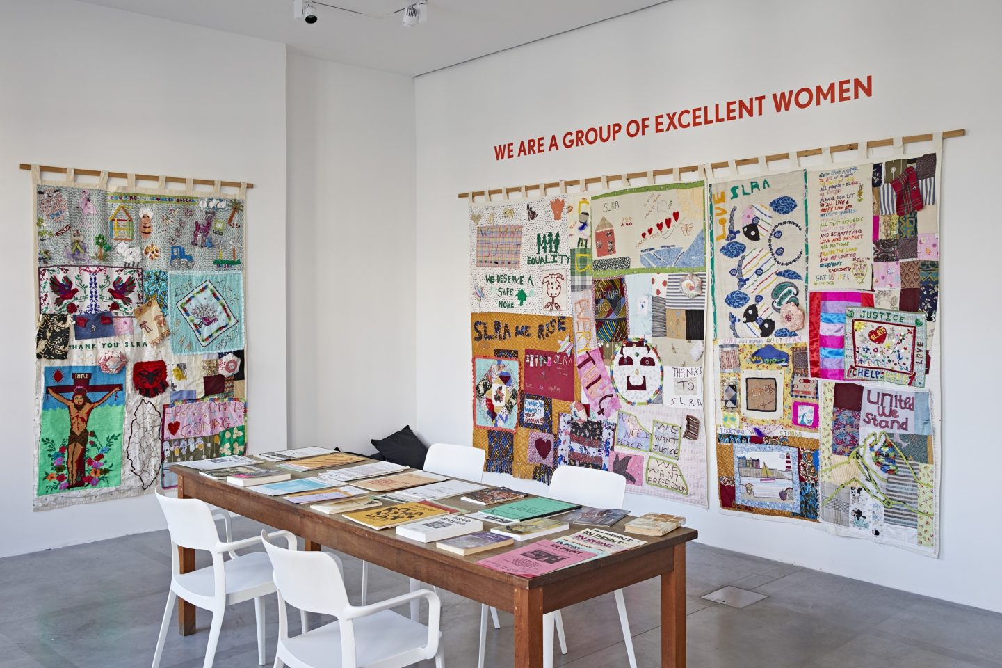 Photo of a gallery space with a table and chairs in the centre and books and other reading material laid out on the table. There are two tapestries with multiple works of art attached to the fabric hung on the wall on the right and left hand side. The exhibition title 'We are a Group of Excellent women' is laid in red on top of the tapestry on the right.
