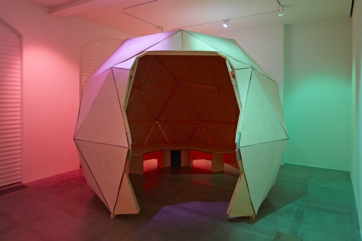 Image of a dome structure in the centre of a gallery space made from large triangular cut-outs. There is an entrance into the dome with a bench inside. There are also spotlights of the colour green on the right-hand side and the colour red on the left-hand side of the dome.