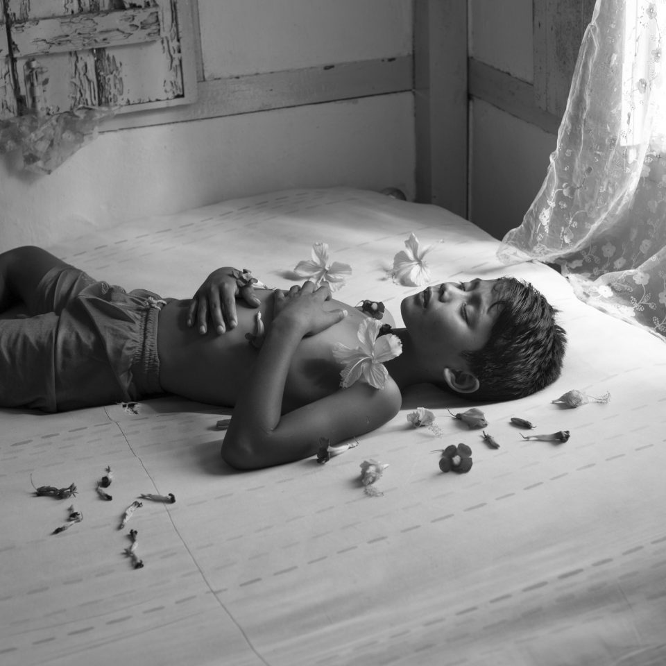 A black and white photo of a topless person lying on a bed with flower petals around them. Their hands are resting on their chest and their eyes are closed. Wooden panellng can be seen on the wall and a sheer curtain drape can be seen on the right-hand side of the window letting in a soft light into the room.