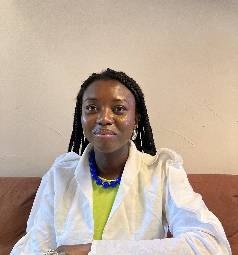 A woman wearing a white shirt, yellow t shirt and blue necklace is sitting down and looking at the camera. She is smiling.