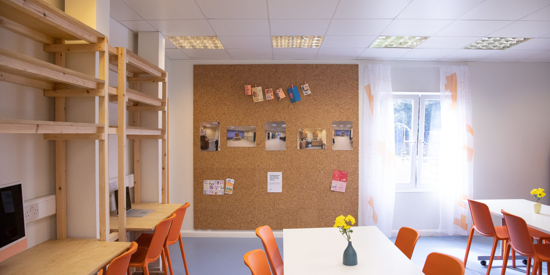 A bright community centre room. There are lots of orange chairs and a big notice board with photos on it. There is a vase with daffodils on the table.