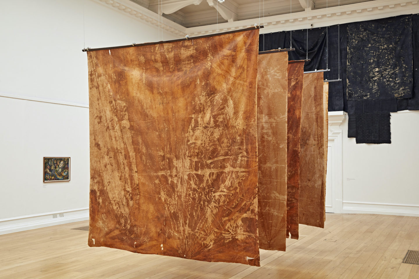 A photo of a large gallery space with multiple art installations. There are five large consecutive orange-coloured drapery in the centre of the gallery space suspended from the air, a small painting on the wall on the left -hand side and a large black draping on the back of the gallery space attached to the wall.