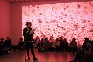 An artist standing up, reading text on in a red room, surrounded by people sitting on the floor. 