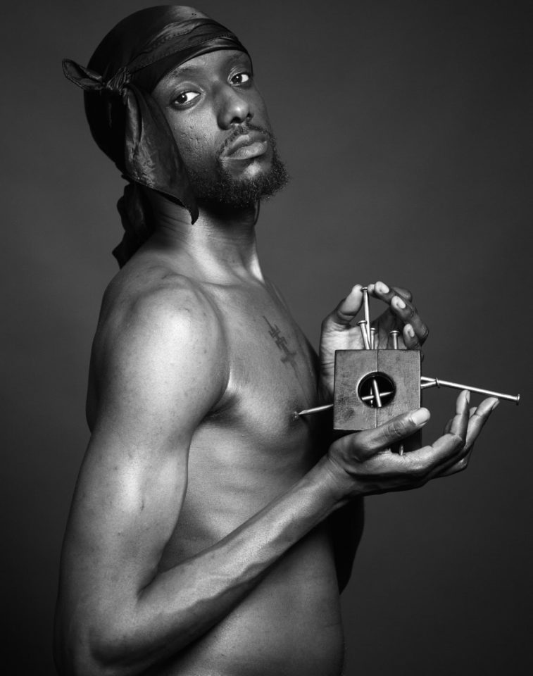 Black and white photograph of a topless man holding a small wooden object