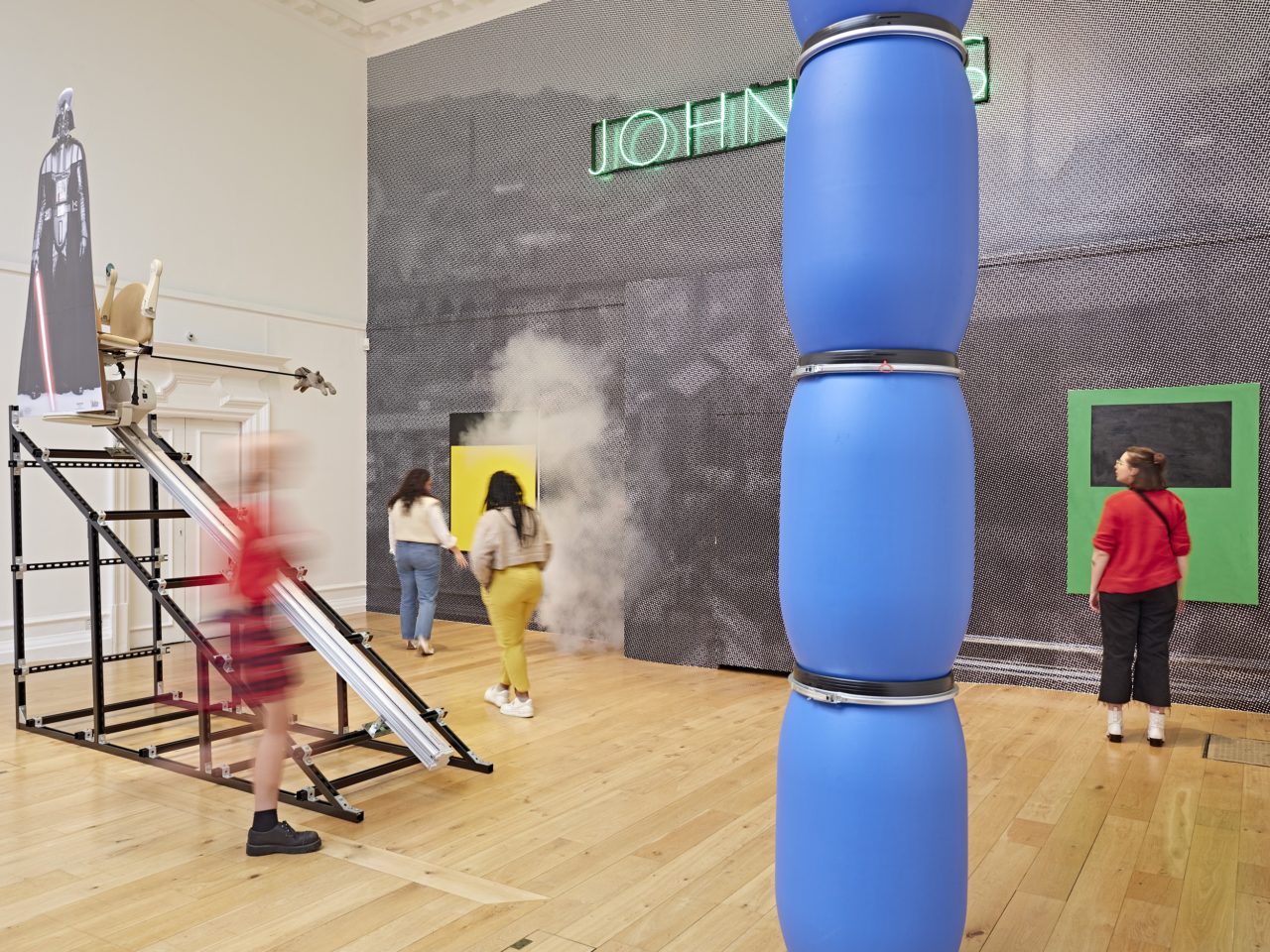 An image of multiple installations in a large gallery space. Blue barrels are stacks in front on top of each other, a cut-out of Darth Vader can be seen on top of an incline structure, a green painting hung up on the wall, a large greyscale wallpaper hung on the wall with a neon green light labelled 'JOHNNY'S' positioned at the top of the wall. There are also several figures walking around the gallery area.