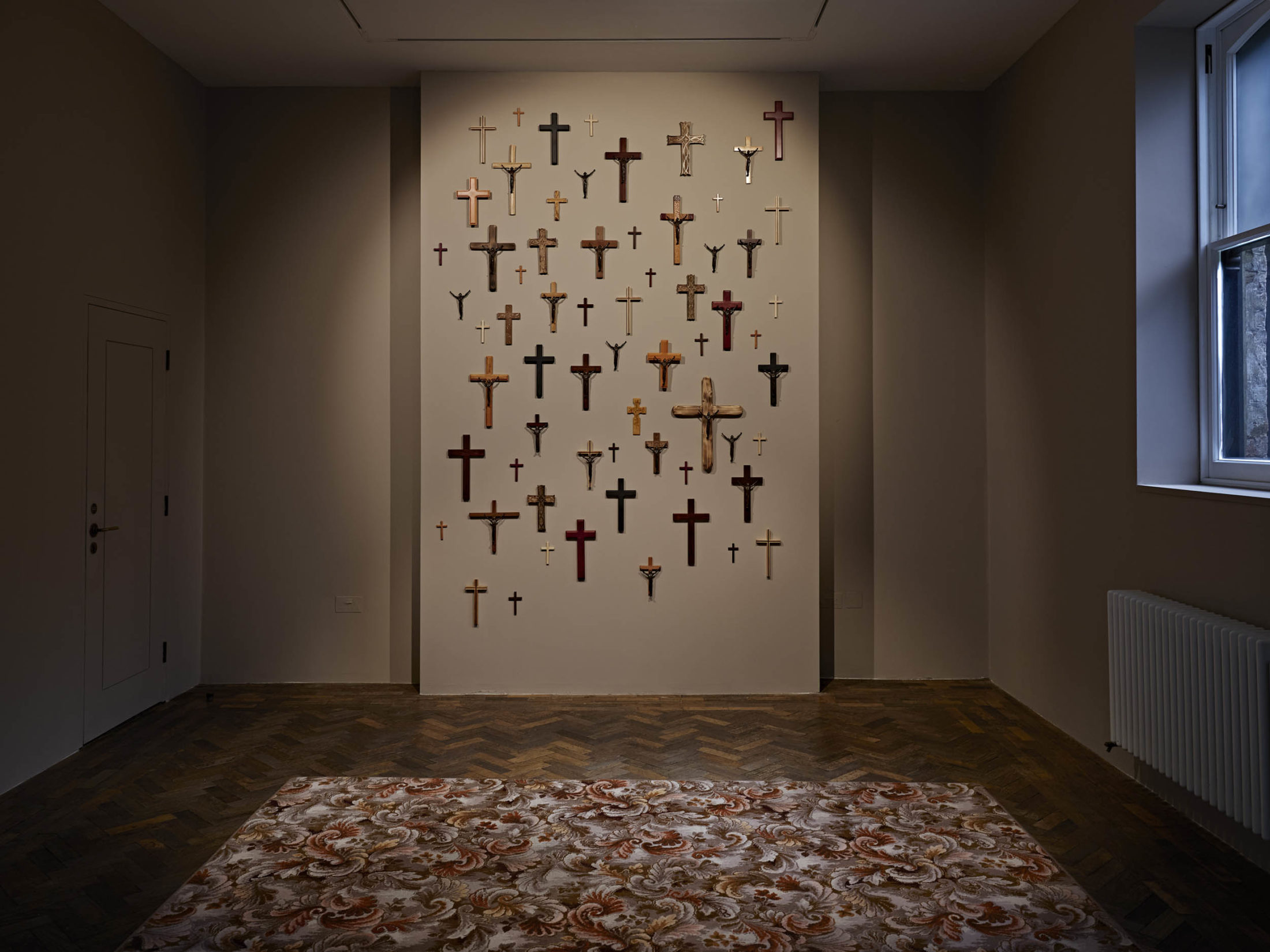 A dark art gallery room. A floral carpet is on the floor and one wall of the gallery is covered with wooden crosses.