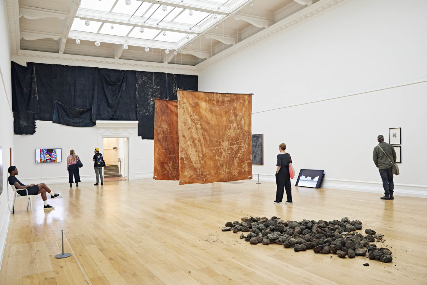 A photo of a large gallery space with multiple art installations. In the middle of the gallery, there are many rock structures are assembled together and there are two large orange drapes suspended from the air. There are also multiple pieces of art work that are framed and hung across the walls of the gallery and digital screens also by the walls of the gallery. Several visitors can be seen viewing the art work across the gallery space.