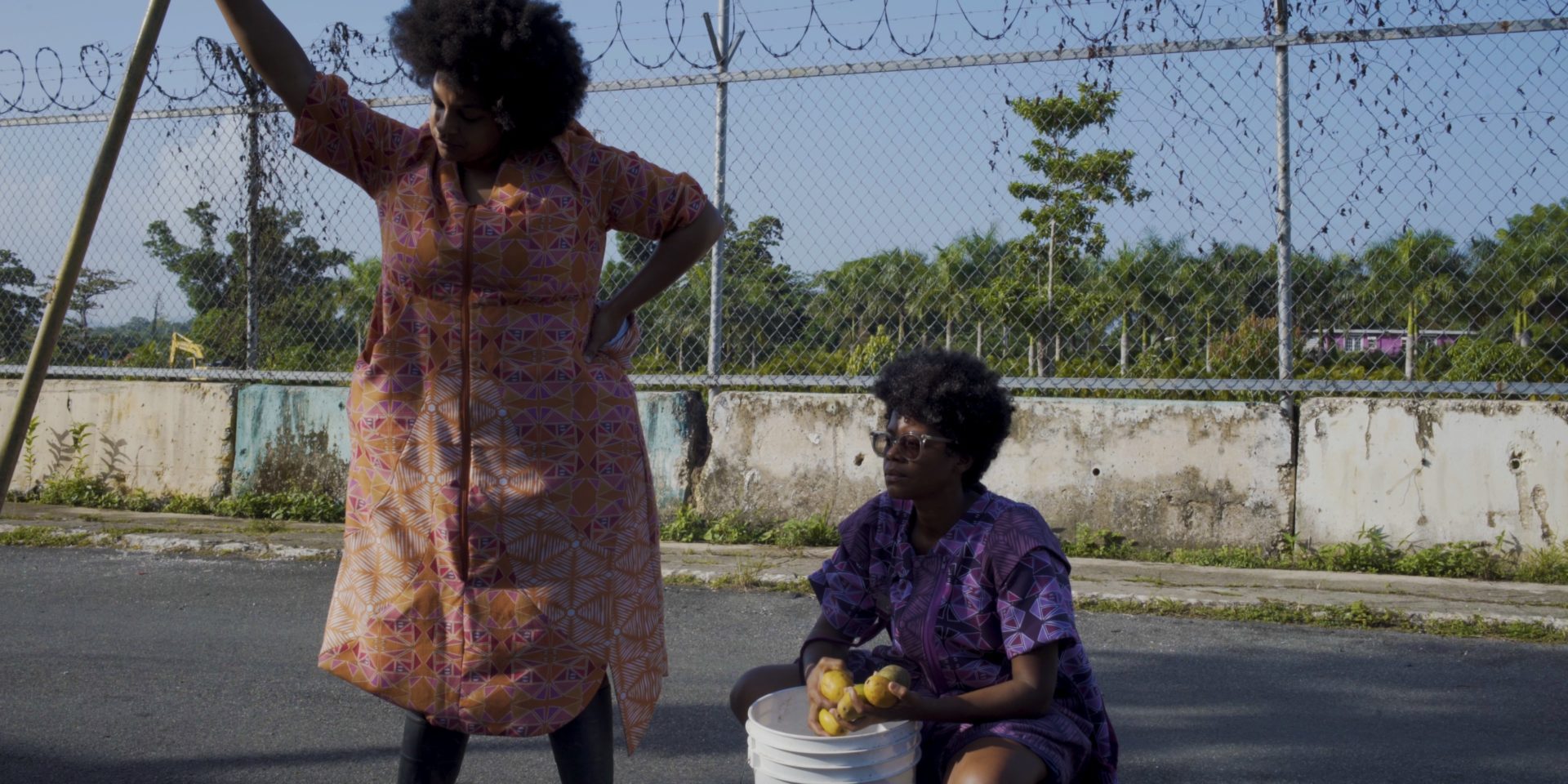 Two people in what looks like a concrete car park outside. One crouches down by a bucket. The other is standing and leaning against a large stick.