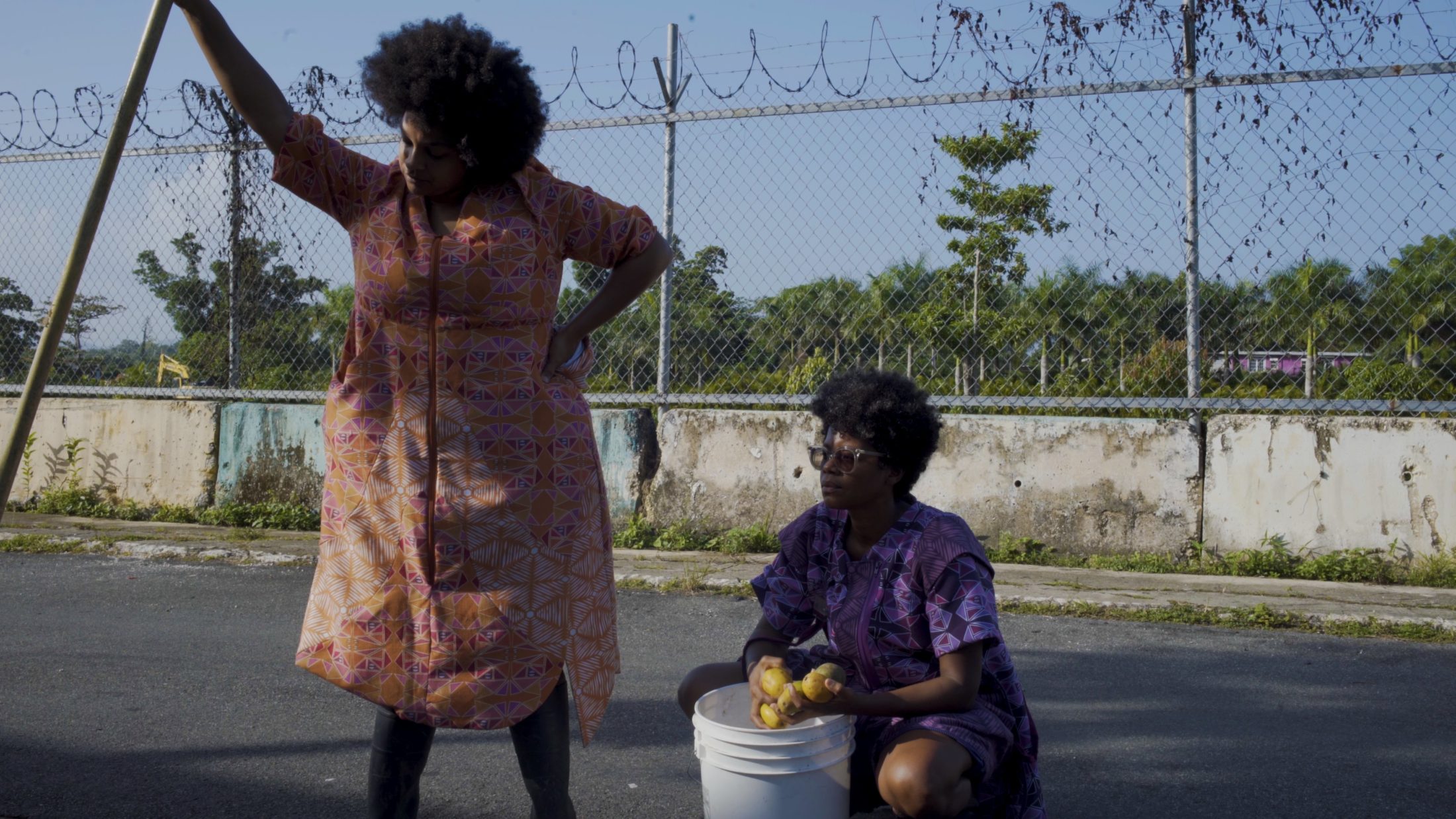 Two people in what looks like a concrete car park outside. One crouches down by a bucket. The other is standing and leaning against a large stick.