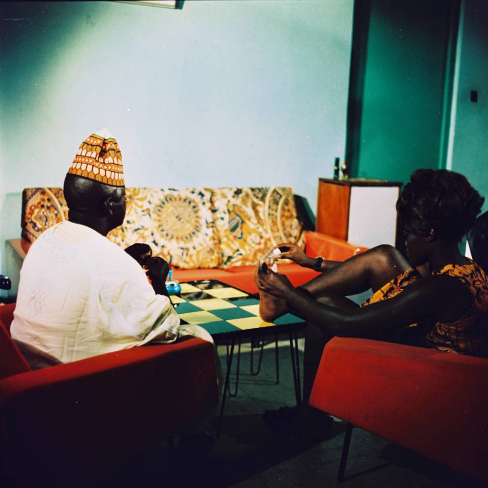 Two people sit on orange armchairs in a living room with their backs to us. The man wears a white shirt and a yellow hat.