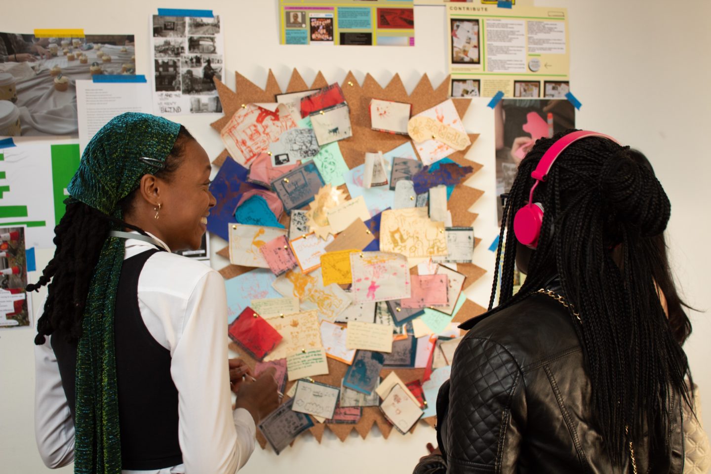 Two young women stand in front of a pin board with lots of paper, post its and colourful notes stuck to it.