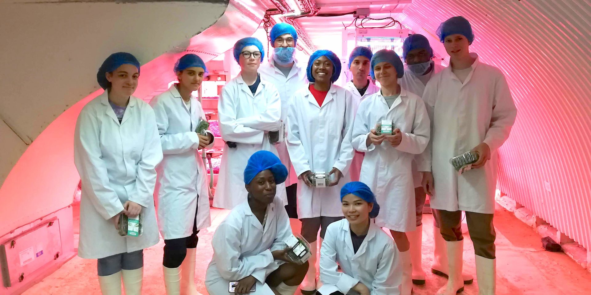A group of young people pose for a group photo inside a underground tunnel lit by a pink light. They are wearing white lab coats and protective nets on their hair and faces.