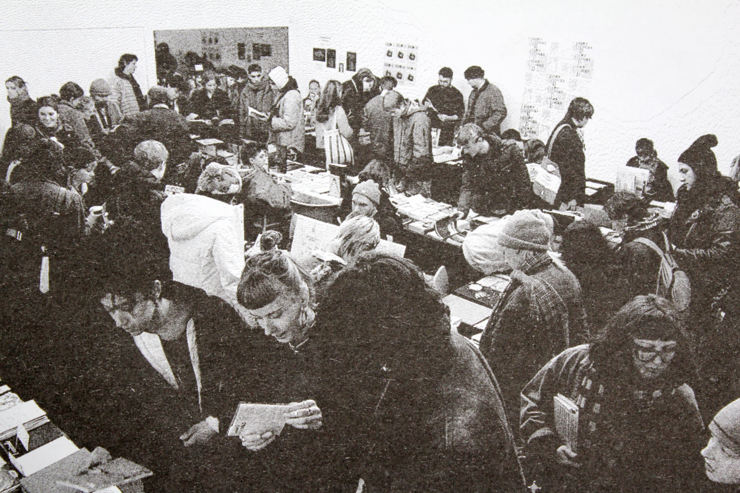 A black and white grainy image of a crowd of people standing around tables holding books and zines