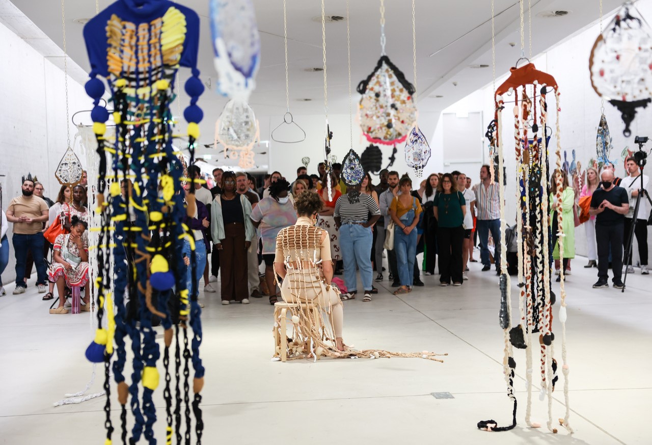 A woman wearing a woven costume sits on a chair in front of a crowd of people in a gallery. Behind her are hanging woven sculptures.