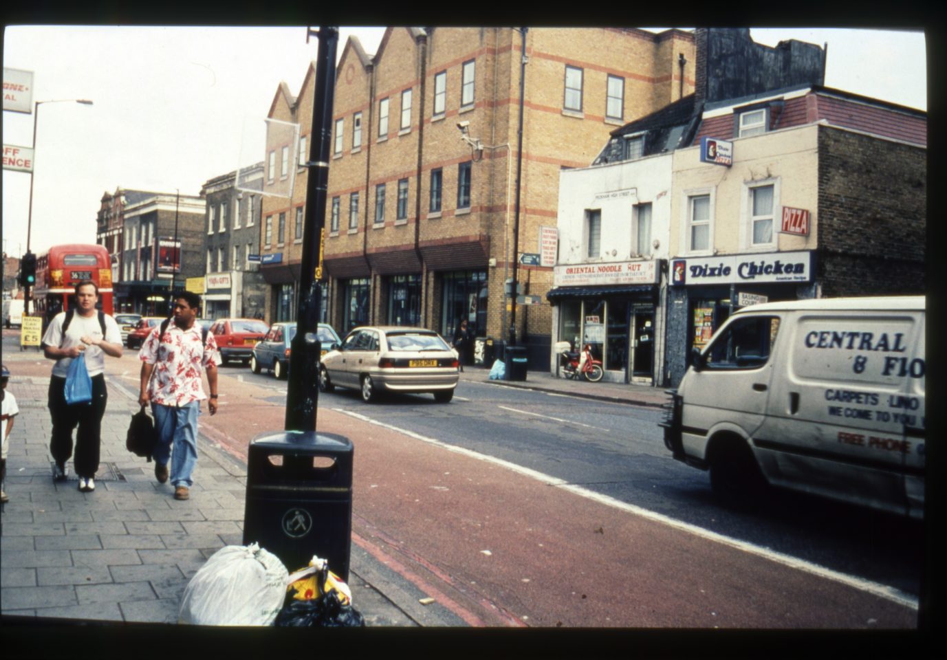 An old photograph of a road in Peckham. Cars and vans drive down the road and two men walk beside each other on the footpath.