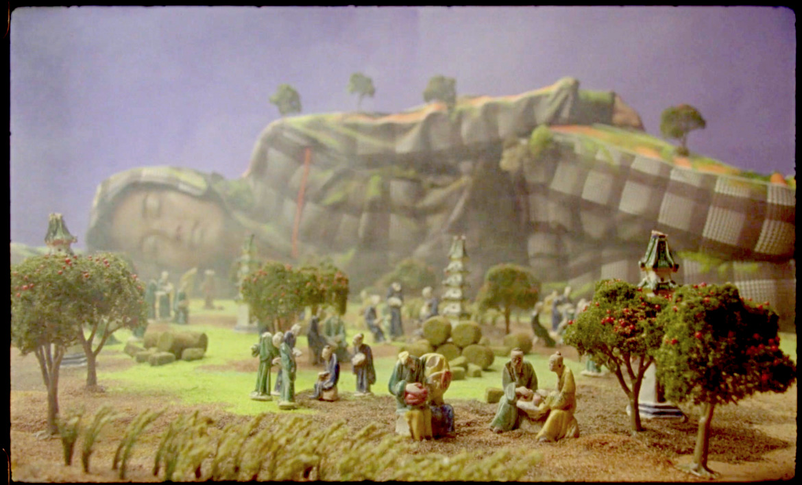 A still from a film with a person lying down in the background and a set of miniature figurines in the foreground.