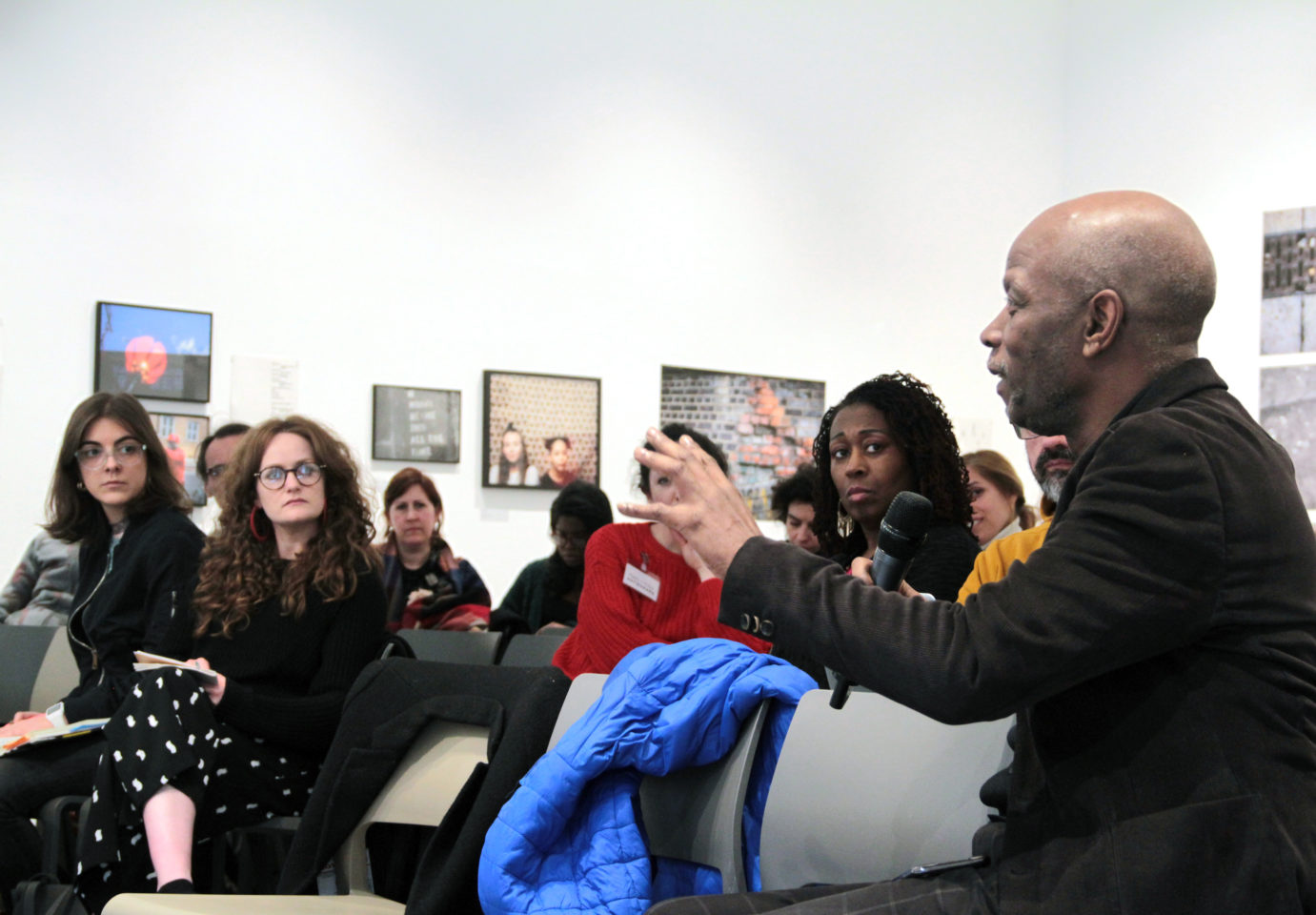 A man holds a microphone and speaks to a room full of people sitting and looking at him