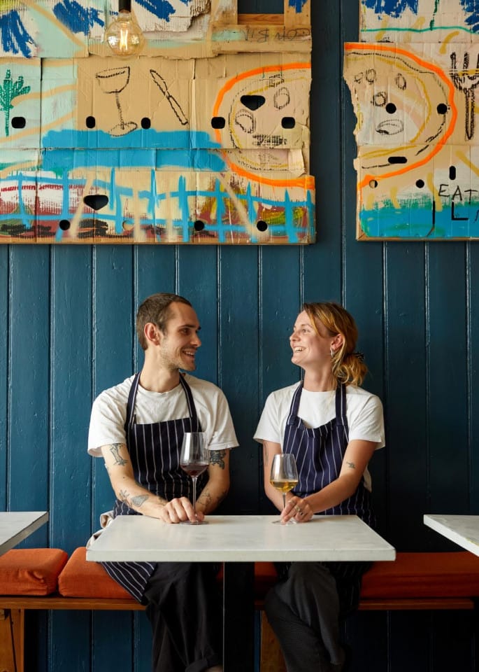 Two chefs sit in a bright colourful restaurant wearing aprons. They are looking at each other and smiling.