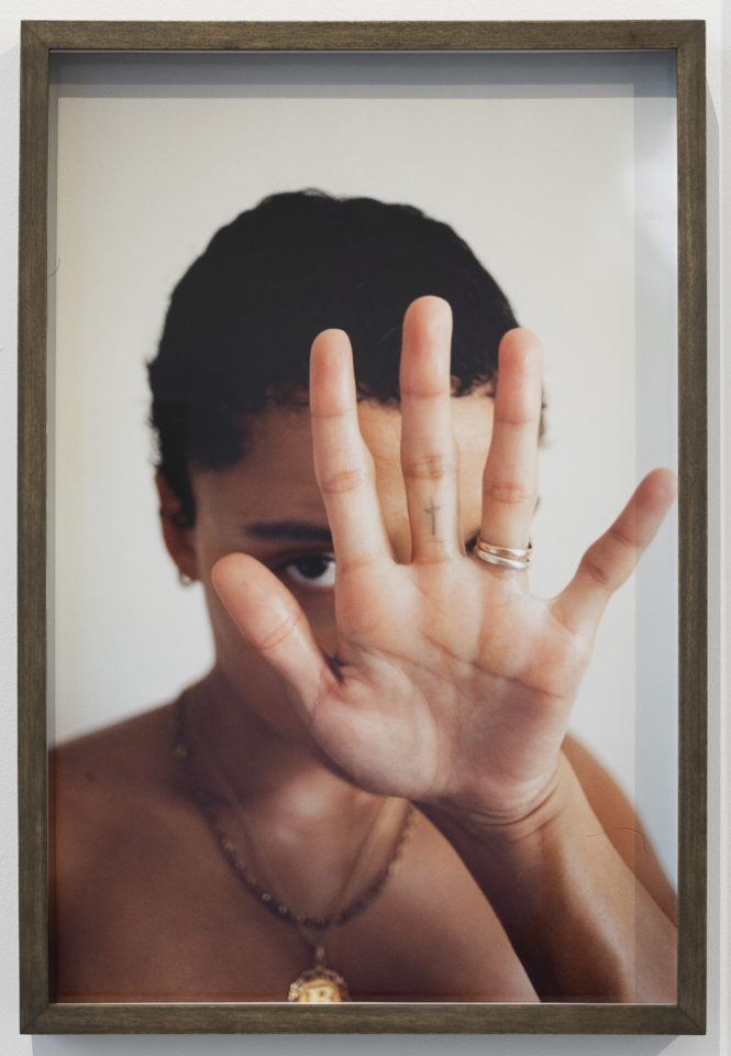 Photograph of the artist Rene Matić cut off just above their chest with a white background. The artist is wearing a pendant necklace and their left hand is spread out towards the camera and covering their face, only their right eye can be seen looking into the camera. Two rings stacks upon themselves are seen on the artist's ring finger and a small cross tattoo is also seen on the base of their middle finger.