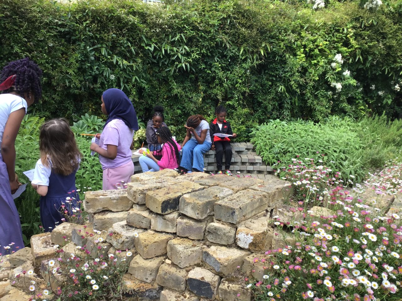 A group of children holding paper worksheets in a garden with a brick feature.
