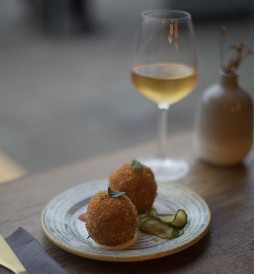 Two fried balls sit on a plate with garnish. A glass of wine is on the table beside them.