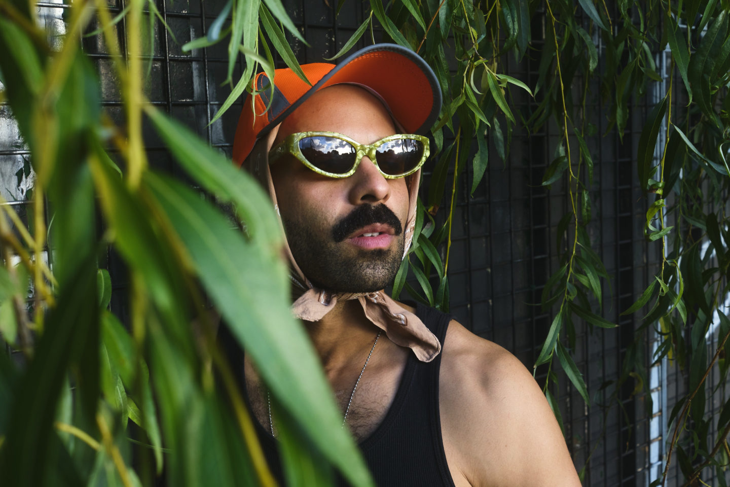 A person wearing an orange cap and sunglasses, with a scarf tied underneath their chin stands amongst green leaves.